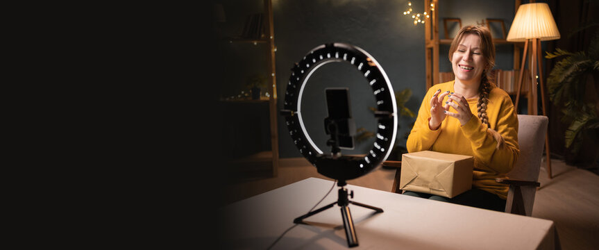 Tricky Female blogger unpacking box recording video using smartphone, ring light with tripod clasping hands and looking away late evening at home office. online shopping and social media