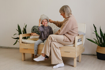 An elderly woman helps a young child put on a knitted hat while sitting on a cozy wooden bench. The room is softly lit with indoor plants around