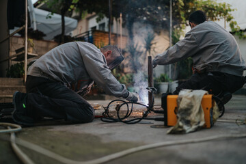 Workers in protective jackets weld metal pieces outdoors, sparks flying as they lean in and focus on the task. Portable welding machine, cables, and safety gear are visible in a street side workshop.