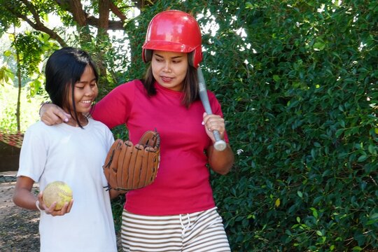 The woman wears a red helmet and holds a baseball bat, with her arm around the smiling girl who is holding a ball.