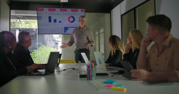Male manager presenting data to diverse business team in modern conference room while screen displays charts and colleagues listen around meeting table - Powered by Adobe