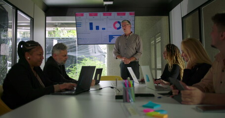 Male manager presenting data to diverse business team in modern conference room while screen displays charts and colleagues listen around meeting table