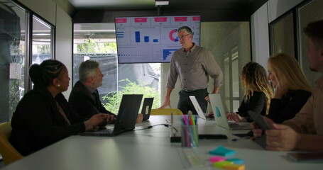 Male manager presenting data to diverse business team in modern conference room while screen displays charts and colleagues listen around meeting table