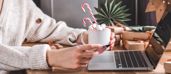 Online Christmas shopping with credit card and laptop, gift boxes and candy cane mug on desk —...