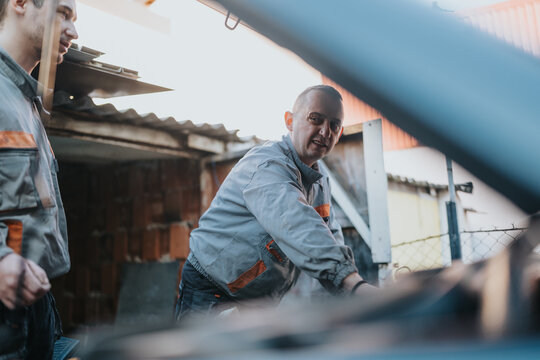 Two mechanics in gray uniforms work to inspect and repair a vehicle under the open hood. A collaborative moment outside a brick workshop conveys hands-on service and teamwork.