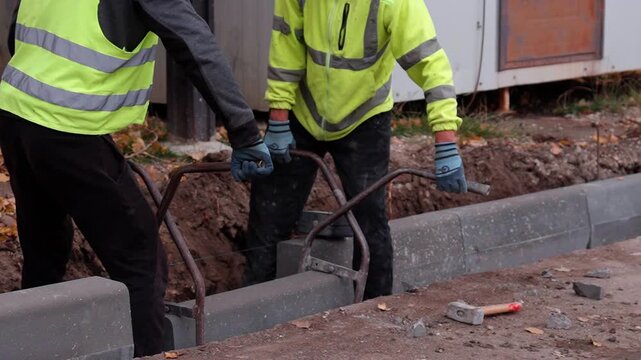 Construction workers in safety gear are installing concrete curbs along a roadway, showcasing teamwork and precision, with a focus on the lifting action and equipment used