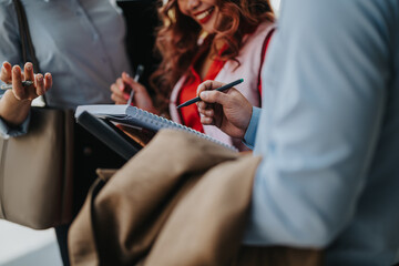 A group of people gathers outside, exchanging ideas with notebooks and pens in hand. They...