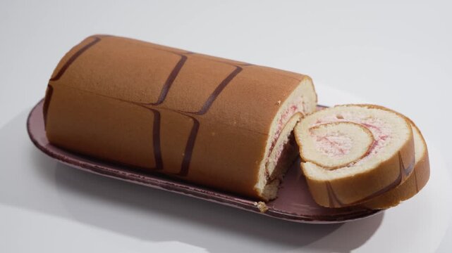 Studio video of a sponge cake roll with chocolate stripes and a strawberry cream spiral on a dark plate. The dessert rotates smoothly on a white background.