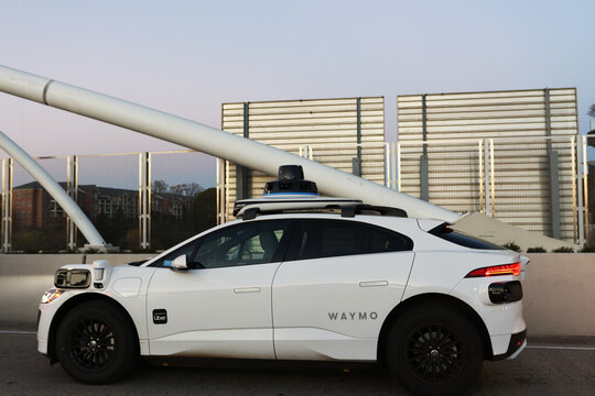 A sleek white autonomous vehicle branded with Waymo is captured in a clean side profile against an urban backdrop at dusk. The car features prominent rooftop lidar sensors and side-mounted camera unit