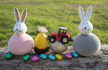 Two souvenir Easter bunnies, toy model of a tractor standing on painted egg, with many multicolored chocolate eggs nearby. Concept for commercial Easter greeting for farms, agribusinesses. Postcard.