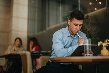 A man in a light blue shirt concentrates on a tablet with a stylus in a modern cafe. Background figures chat, while a yellow vase and flowers add a warm, casual business vibe.