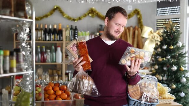 Interested young man standing with shopping basket among holiday decorations in grocery shop, holding freshly baked panettone, marzipan cookies and nutty nougat, choosing desserts for Christmas feast