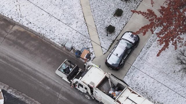 Drone shot looking down from aerial above garbage truck driving along neighborhood street and collecting trash, waste, and can junk by side of homes and house yards with claw during cold snowy winter