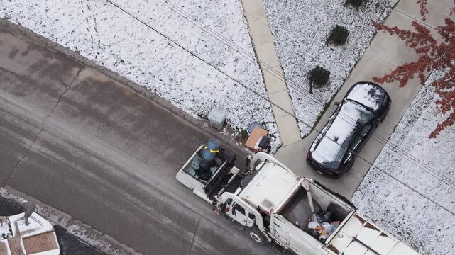 Drone shot looking down from aerial above garbage truck driving along neighborhood street and collecting trash, waste, and can junk by side of homes and house yards with claw during cold snowy winter