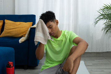 Brown skinned teenager sitting on an exercise mat in his living room, wiping off sweat after working out. Image of effort, well being, and home training