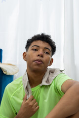 Dark skinned teenager in a close up shot, sweating and looking tired while resting seated on an exercise mat with a towel around his neck. Home workout routine