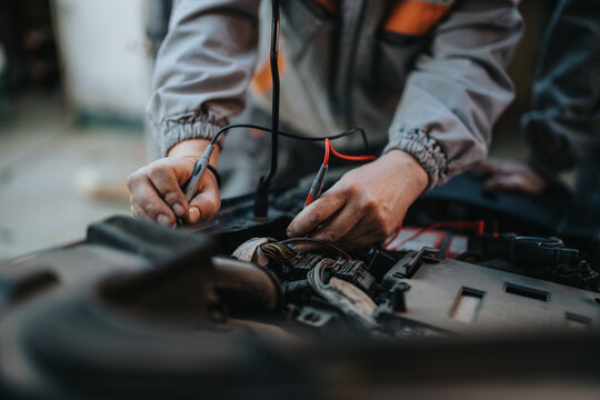 A mechanic works on a vehicles electrical wiring, using probes to test circuits in the crowded engine bay. Focus on hands, tools, and orange work wear conveys skilled auto repair.