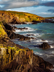 A breathtaking view of the rugged Irish coastline. Ocean waves crash against the rocks below the cliffs. The sky is blue with scattered clouds, showing a beautiful landscape.