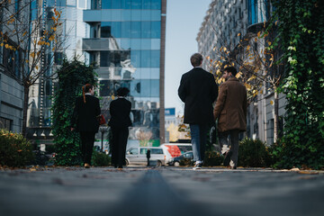 A group of business people in coats stroll along a modern urban street, autumn leaves scattered as glass towers and ivy create a vibrant city scene.