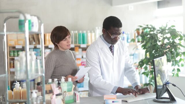 African man pharmacist uses computer at workplace and consulting woman client. Staff reads instructions on computer, interprets medical prescription and makes list of drugs to sell to client