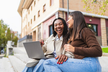 Two diverse women friends enjoying a happy moment, smiling and using a laptop while sitting on steps outside a building