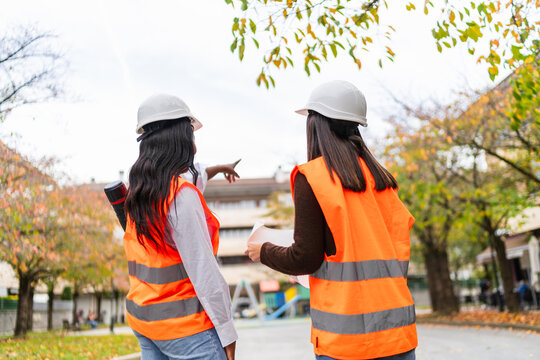 Two diverse women engineers in hard hats and safety vests discussing a construction plan, pointing at a building structure