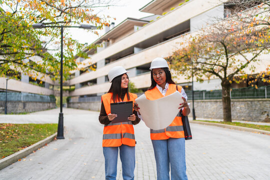 Two women engineers wearing hard hats and safety vests are analyzing a blueprint while working on a construction project