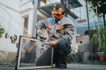 A laborer in protective goggles grinds a glass pane with a handheld grinder in an outdoor workspace. The scene captures focus, skill, and practical construction activity.