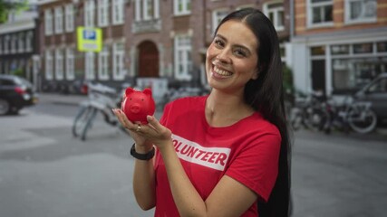 Woman holds red piggy bank in hands on street smiling and holding it forward as donation gesture; community fundraising generosity.