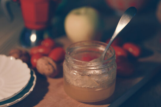 Close-up of a jar of honey with a teaspoon; fruit and various other items on the table.