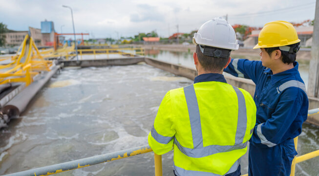 Two professional male engineers discussing maintenance plans on a clipboard while standing on a metal walkway at a large industrial water treatment plant and sewage facility outdoors.