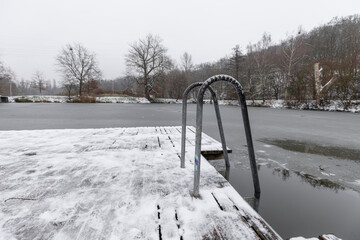 Snow covers a wooden pier with metal handrails entering a partially frozen pond in winter. Horejsi rybnik in prague 9