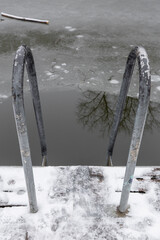 Fototapeta premium Metal ladder handrails entering a partially frozen water pond with snow covered deck. Horejsi rybnik in prague 9