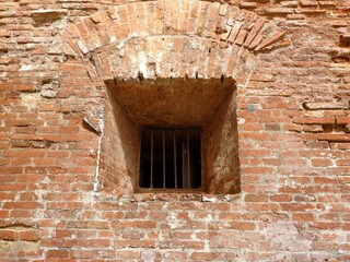 Textured, weathered red brick wall with an arched window opening secured by rusty, vertical metal bars. A classic, historic architectural detail suggesting age and confinement.