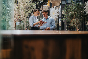 Obraz premium Two colleagues in light blue shirts discuss ideas while looking at a tablet in a stylish indoor space. Greenery and decor create a calm, collaborative, modern atmosphere.