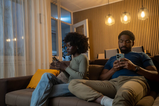 Young African American couple sitting apart on couch at home, using phones separately
