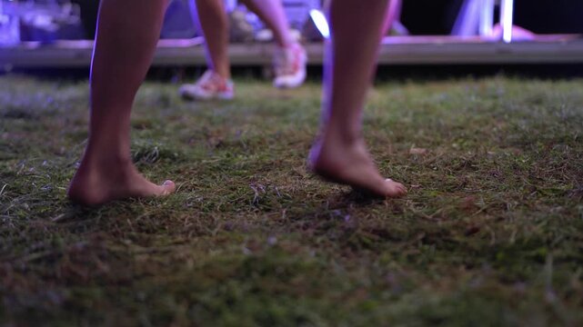 People dancing at an outdoor music festival on a summer night. A close-up of a girl's bare feet dancing to music on the grass. Legs of dancing people.