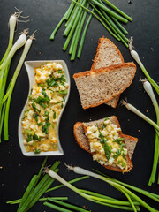 Delicious boiled egg mayonnaise salad sandwich with green leek onion, chopped and mixed yolk and white on crusty bread baguette with portion of salad on black kitchen wooden table, top view.