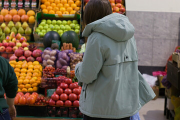 In a vibrant marketplace, a young woman with a blue shopping bag pauses in front of a display of fresh fruits and vegetables. Her attention is fixed on the colorful produce.