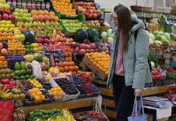 A young woman stands at a fruit and vegetable stall in an indoor market. She holds a bag in her...