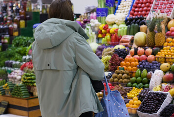 In a vibrant marketplace, a young woman with a blue shopping bag pauses in front of a display of...