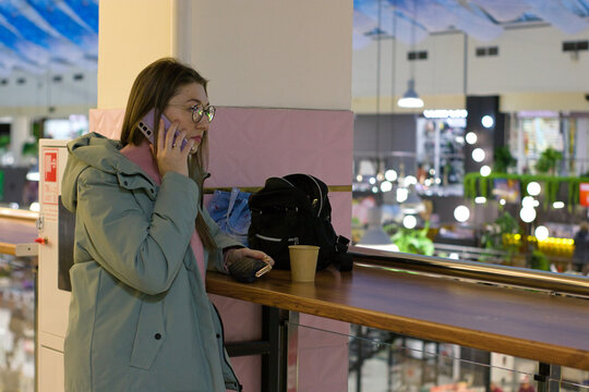 A young woman stands at a counter in a brightly lit public area, holding her phone to her ear. She appears to be in conversation, with a serious expression and a black handbag on the table nearby.