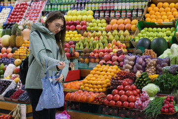 In a vibrant marketplace, a young woman with a blue shopping bag pauses in front of a display of fresh fruits and vegetables. Her attention is fixed on the colorful produce.