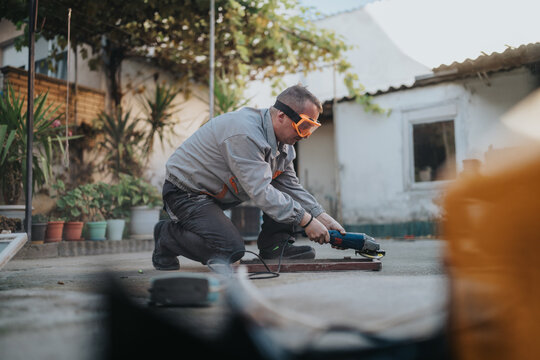 A man wearing orange safety goggles operates a handheld grinder on a work surface in a sunny backyard, surrounded by potted plants and a small workshop area, capturing skilled home improvement.