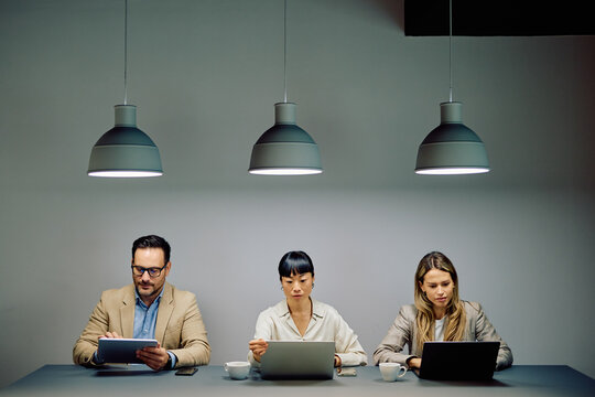 Three diverse business people sitting at a table with laptops and a tablet, focusing on their tasks under pendant lights - Powered by Adobe