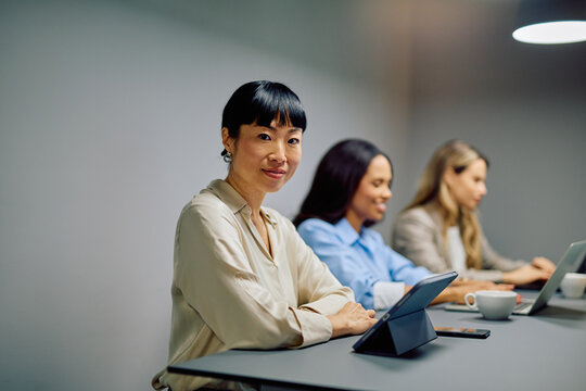 Asian businesswoman smiling at camera, sitting with diverse colleagues at a desk during an office meeting