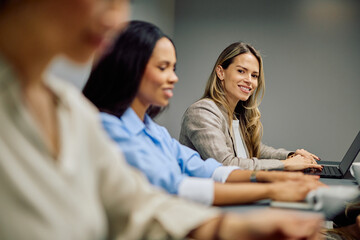 Businesswomen sitting at a conference table, smiling and looking at the camera during a corporate meeting, using laptops