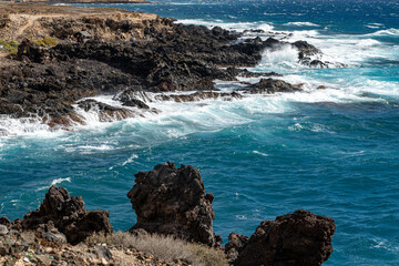 Rocky volcanic coastline of Tenerife with turquoise waves crashing against dark lava cliffs. Dynamic seascape with strong Atlantic wind and dramatic shore.