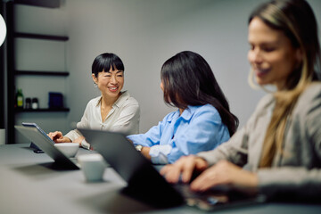 Smiling businesswomen having a discussion while working on laptops and tablets in a modern office, showing teamwork and collaboration