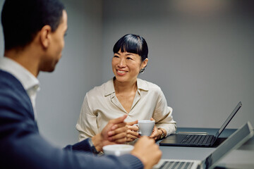 Two diverse colleagues discussing work in an office setting, smiling and enjoying coffee during a professional conversation